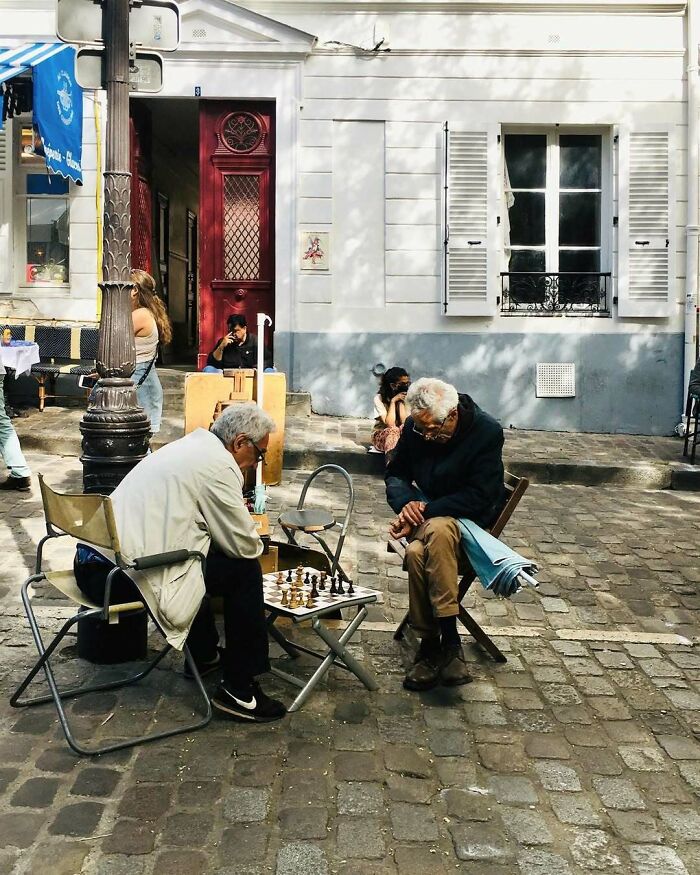 Two stylish grandparents playing chess outdoors on a cobblestone street near a café on a sunny day.