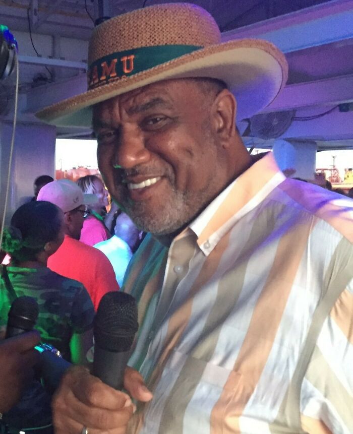 Man in a straw hat holding a microphone, smiling at an event with a crowd in the background during halftime performance.