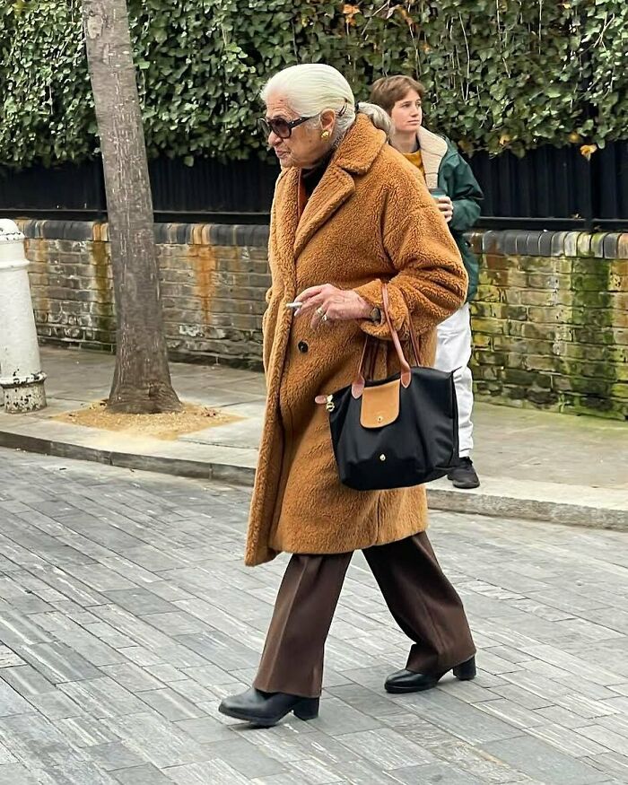 Stylish grandparent wearing a brown teddy coat and sunglasses, carrying a black handbag while walking on a city street.