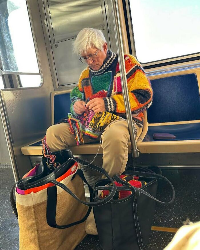 Stylish grandparent wearing a colorful patchwork sweater knitting while sitting on public transit with bags nearby.