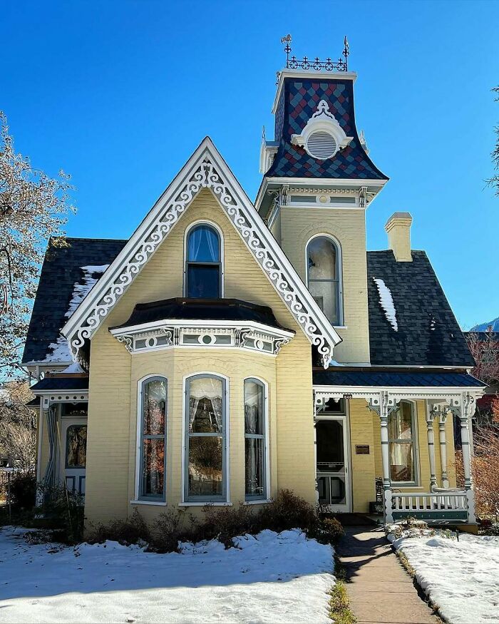 Victorian old home with intricate trim and tall tower under clear blue sky, surrounded by snow and bare trees.