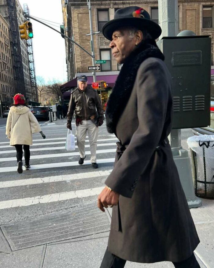 Stylish grandparent walking on a city street wearing a black hat with feather and a long dark coat with fur collar.