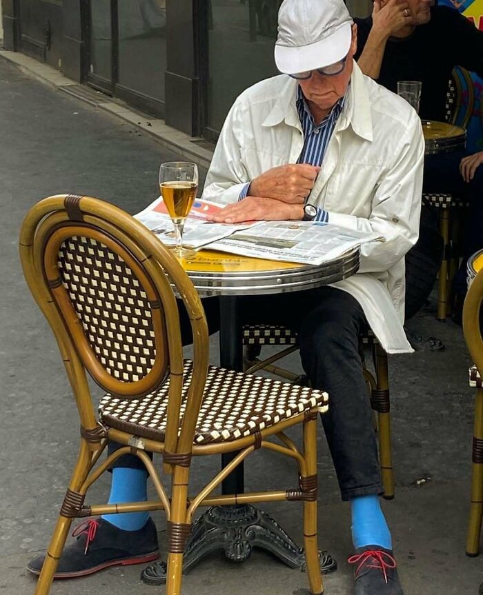 Stylish grandparent in white jacket and cap, wearing bright blue socks and red-laced shoes, sitting at outdoor café reading newspaper.
