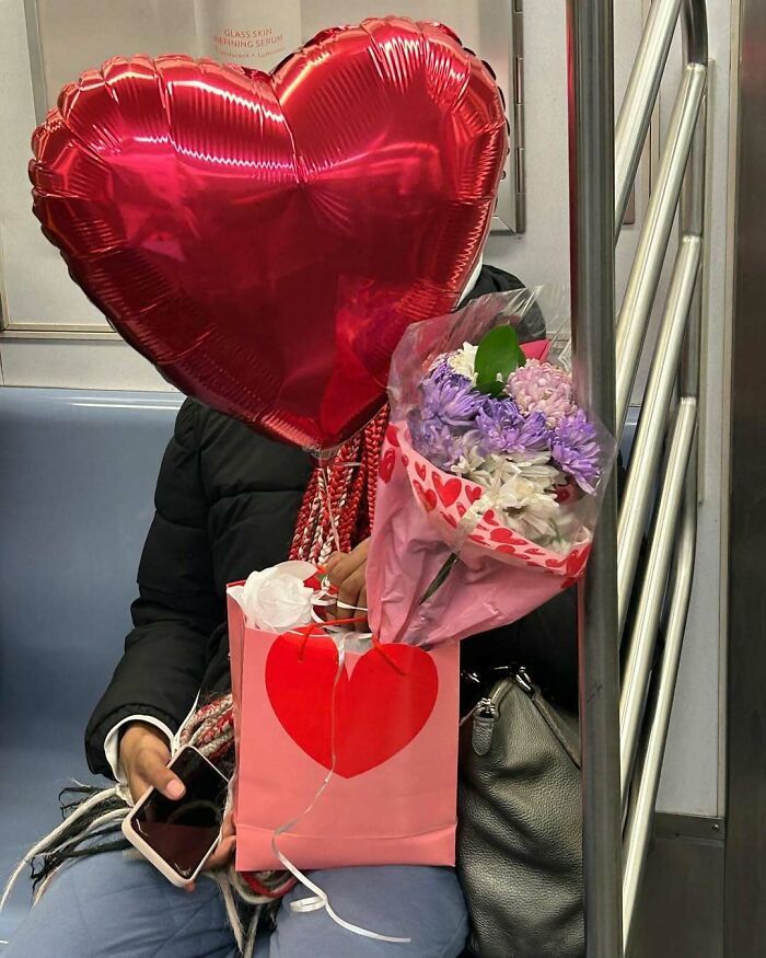 Person on New York subway holding a heart-shaped balloon, flowers, and a gift bag with heart designs.