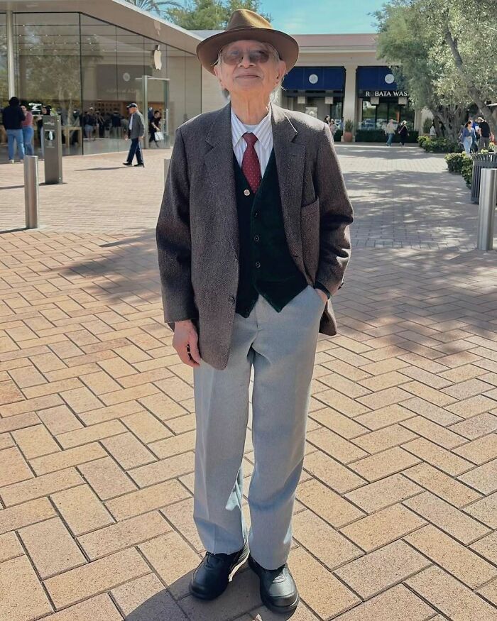 Stylish grandparent wearing a brown hat, gray blazer, red tie, and light gray pants standing outdoors in a shopping area.