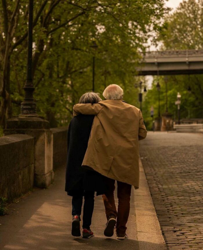 Elderly couple wearing stylish coats walking arm in arm on a tree-lined city street in autumn.
