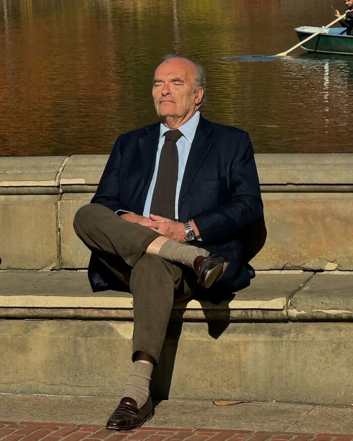 Stylish grandparent sitting by the water in a navy blazer, tie, and brown trousers, showcasing cool and fashionable outfits.