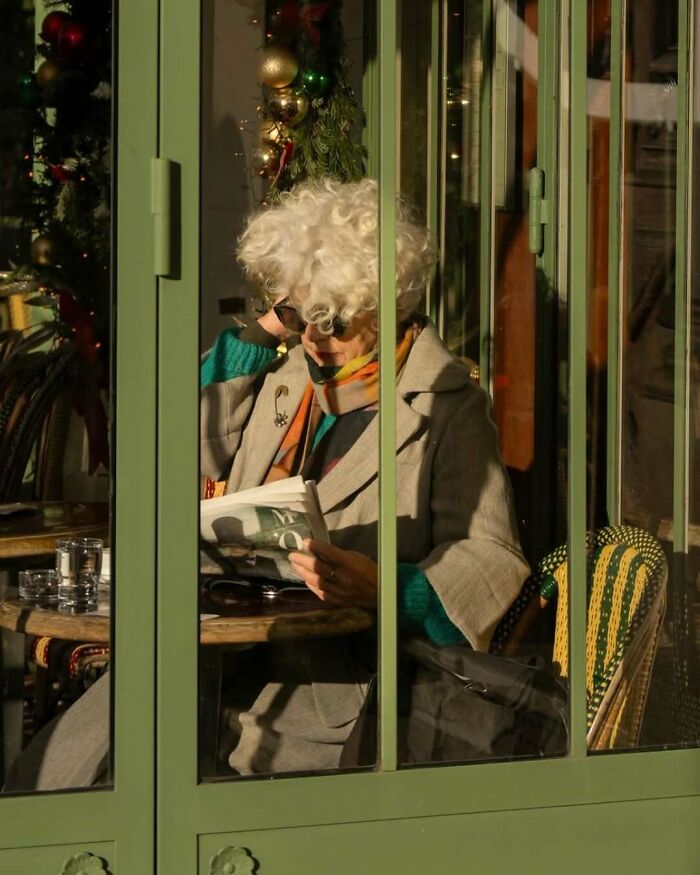 Stylish grandparent with curly white hair reading a newspaper at a café wearing a gray coat and colorful scarf.