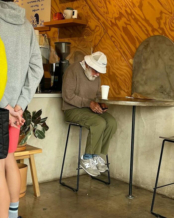 Stylish grandparent wearing a beige sweater, olive pants, white sneakers, and a white bucket hat sitting in a cafe.
