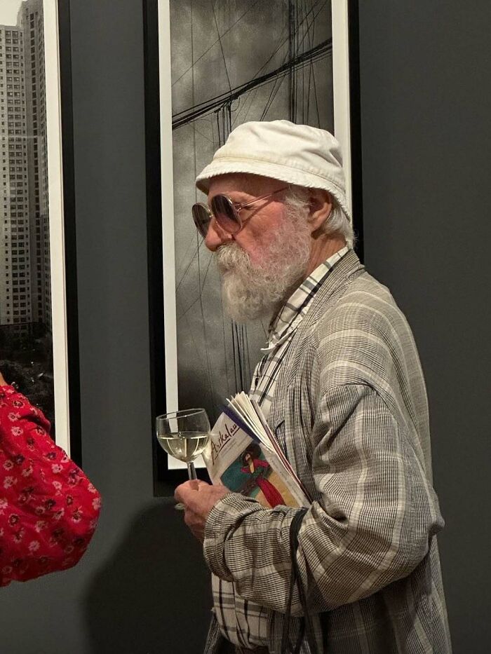 Stylish grandfather wearing a bucket hat and sunglasses, holding a glass of wine and a colorful magazine at an art gallery.