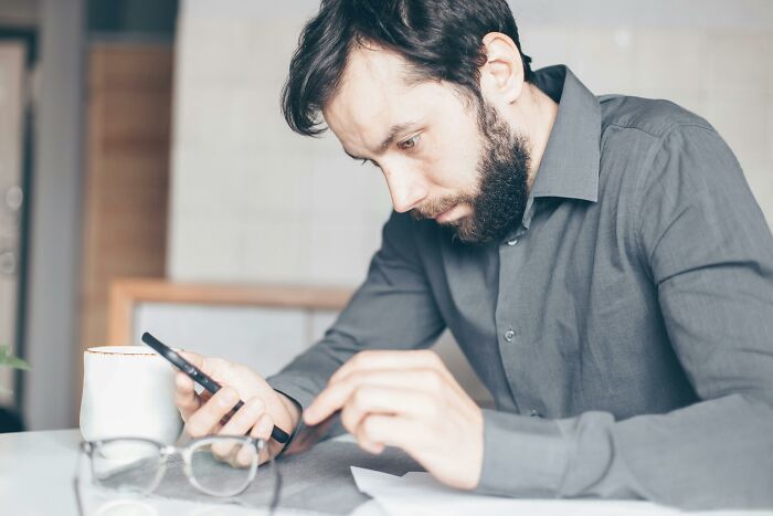 Man in gray shirt drawing the line while focused on his phone and papers at a bright workspace with glasses nearby