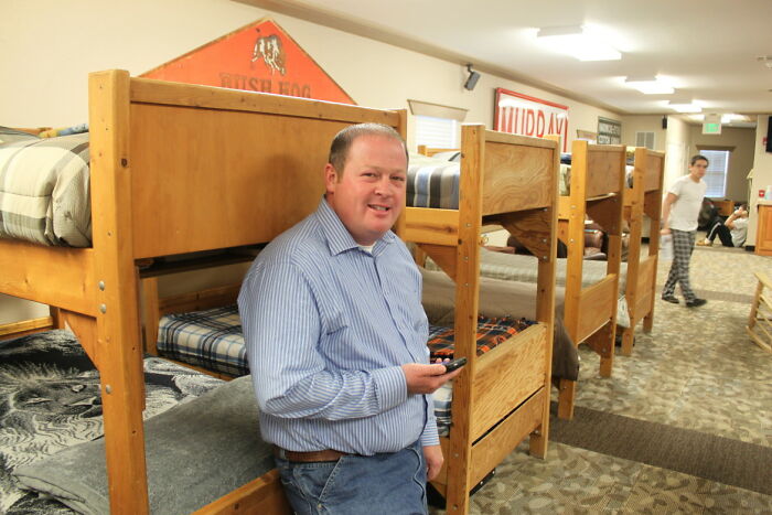 Man in a striped shirt smiling, holding a phone, standing by wooden bunk beds inside a dorm room showing life-changing sentences.