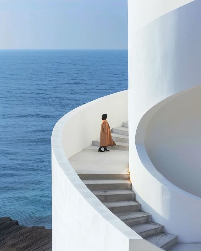 Minimalist photo of a person in a brown coat on white stairs overlooking a calm blue ocean, evoking peaceful escape.