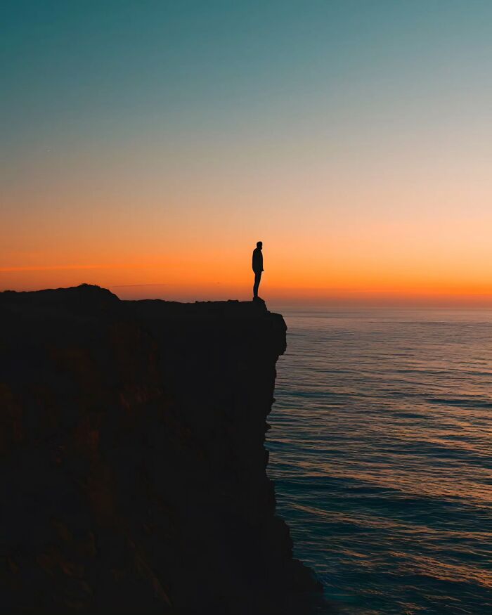 Silhouette of a person standing on a cliff at sunset over the ocean in a dreamy minimalist photo with peaceful tones.