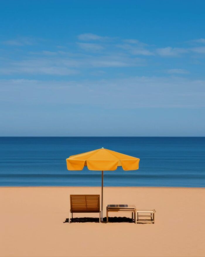 Minimalist beach scene with a yellow umbrella, wooden bench, and table by calm blue ocean under clear sky.