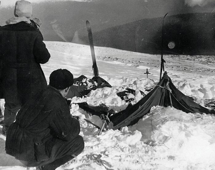 Two people near a small snow-covered tent and skis in a snowy landscape, revealing a mysterious scene.