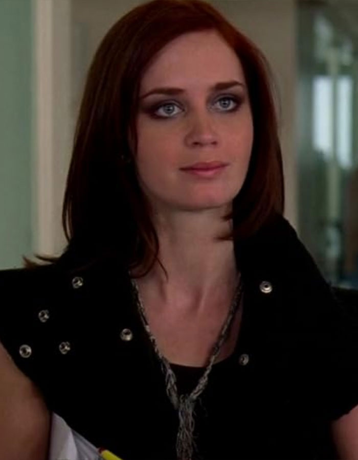 Emily Blunt with red hair and smoky eye makeup, wearing a black studded outfit indoors.