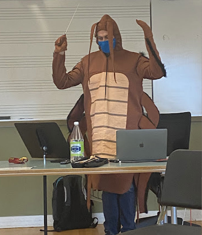 Teacher in a funny insect costume conducting a class, using a laptop and music stand in a humorous teaching moment.