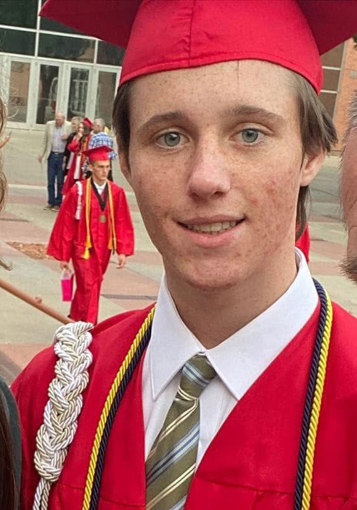 Young man in red graduation gown and cap, wearing honor cords, attending a graduation ceremony outdoors.