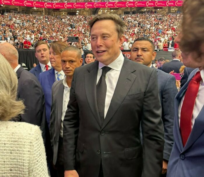 Elon Musk in a black suit at a crowded event, surrounded by people, sparking conspiracy theories online.