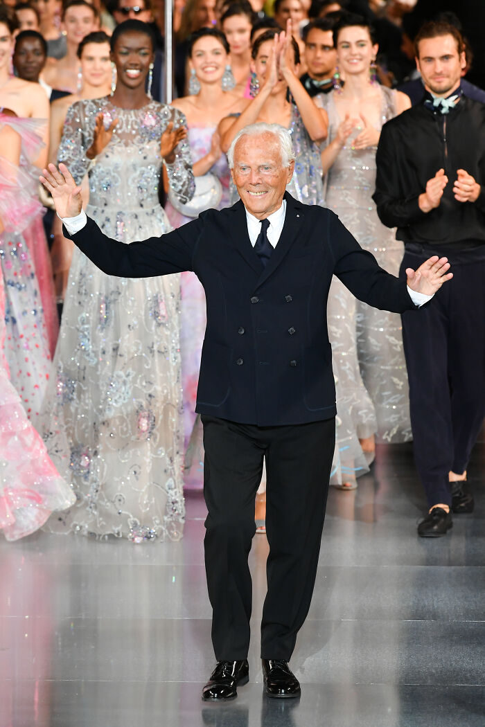 Giorgio Armani, legendary fashion designer, waving and smiling on runway with models in elegant dresses applauding behind him. Giorgio Armani, legendary fashion designer, waving and smiling on runway with models in elegant dresses applauding behind him.