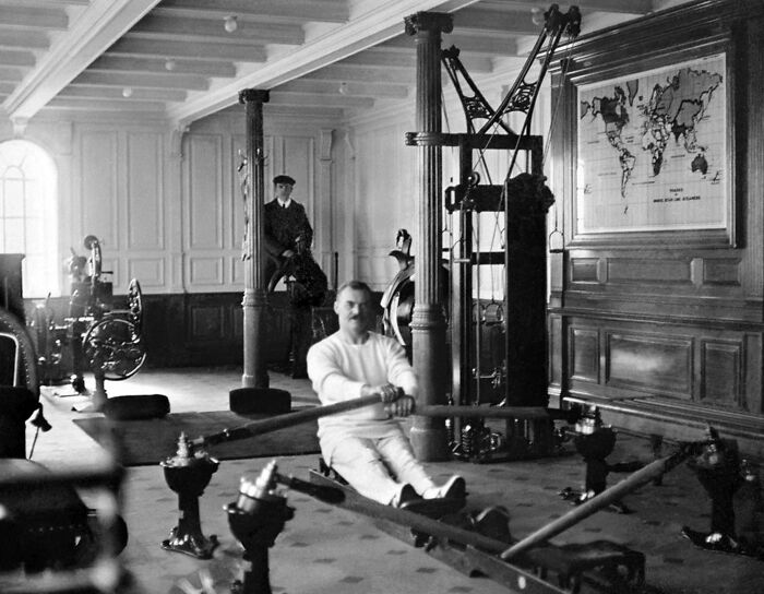 Man rowing on exercise equipment inside Titanic with crew member sitting against column in historical setting.