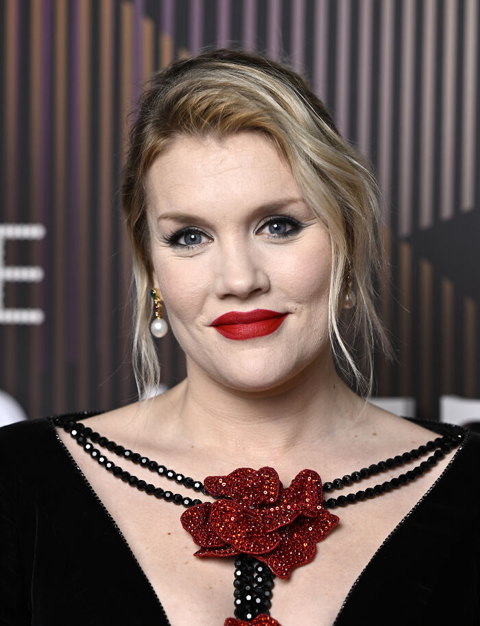 Blonde woman with red lipstick wearing a black dress with red floral embellishment, related to 1800s Wuthering Heights theme.
