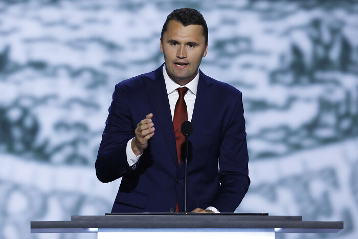 Charlie Kirk speaking at a podium in a dark suit and red tie during a formal event with a blurred background. Charlie Kirk speaking at a podium in a dark suit and red tie during a formal event with a blurred background.