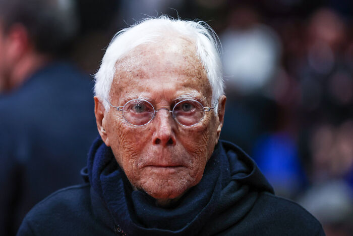 Giorgio Armani wearing glasses and a dark scarf, captured in a close-up portrait with a blurred background. Giorgio Armani wearing glasses and a dark scarf, captured in a close-up portrait with a blurred background.