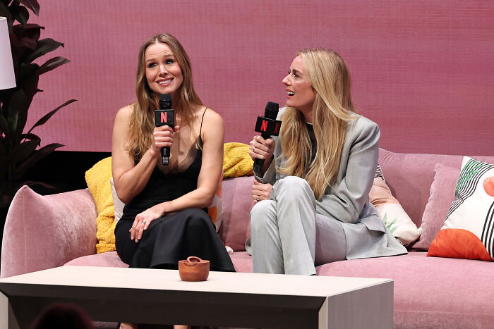 Two women holding microphones on a pink couch discussing biohacking tricks for fitness during a live event.