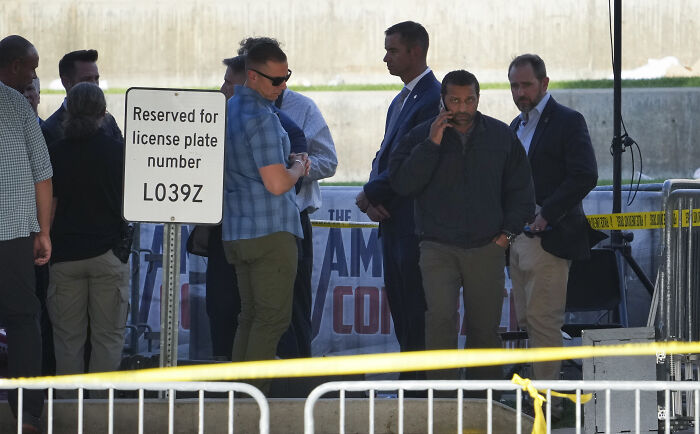 Men in casual and formal attire stand near a reserved parking sign amid FBI security tape in an ongoing investigation scene.
