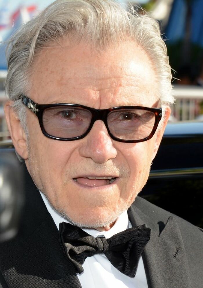 Older man in a black tuxedo and glasses, captured outdoors during a celebrity encounter with a surprising expression.