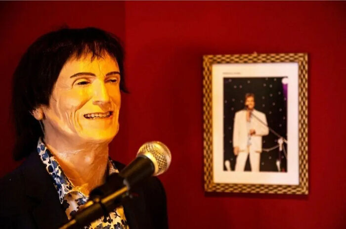 Wax sculpture of a famous figure with dark hair standing at a microphone, showcasing creepy and hilarious wax art by the artist.