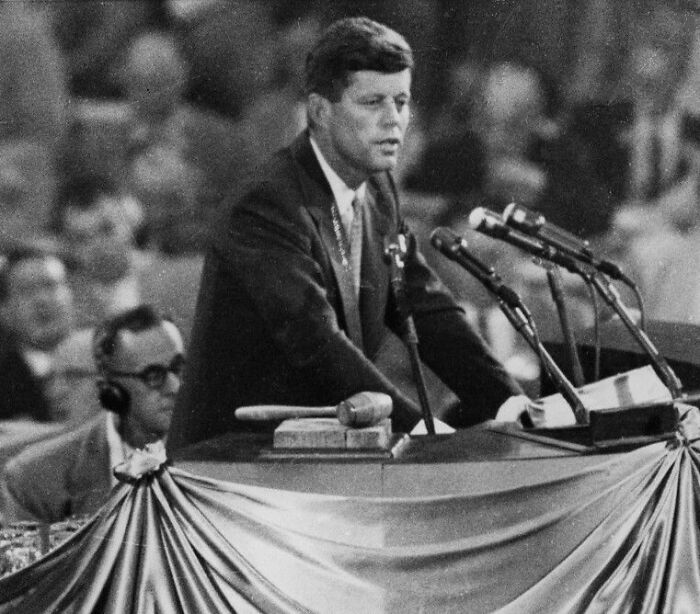 John F. Kennedy speaking at a podium with microphones during a formal event, capturing mass fury comparisons.