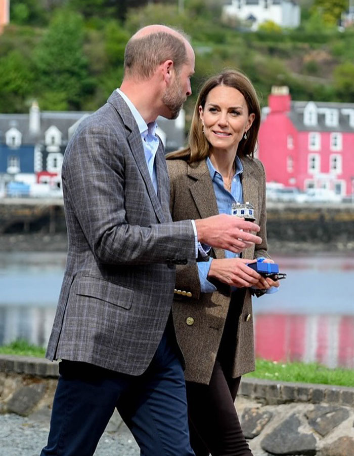 Kate Middleton with brunette hair walking outdoors, holding small objects, with colorful buildings in the background. Kate Middleton with brunette hair walking outdoors, holding small objects, with colorful buildings in the background.