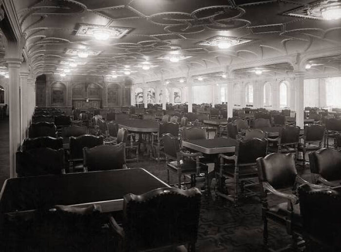Spacious dining area on the Titanic with rows of tables and chairs under ornate ceiling lighting in a historical setting.