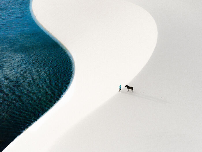 Person standing with a horse on a smooth white sand dune near dark blue water, showcasing minimalism in nature.