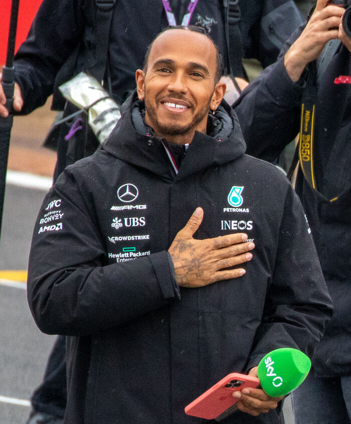 Lewis Hamilton wearing a black jacket with sponsor logos, holding a phone and a Sky microphone in a sports setting.