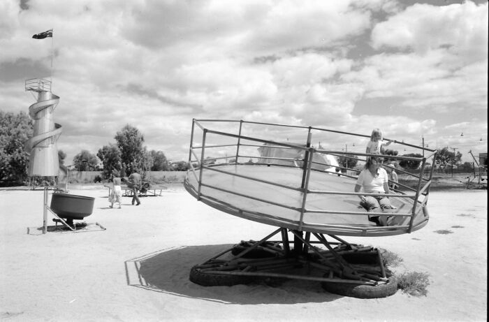 Historic playground equipment with children playing on risky vintage rides under a cloudy sky in a sandy outdoor area.
