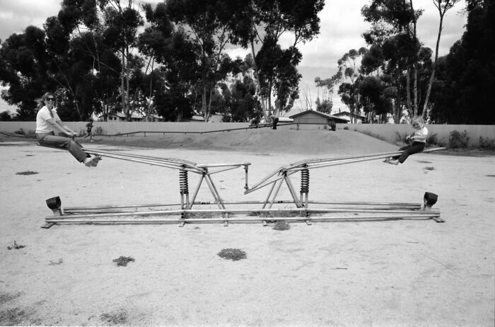 Black and white photo of children playing on a vintage playground seesaw with metal springs and minimal safety features outdoors.