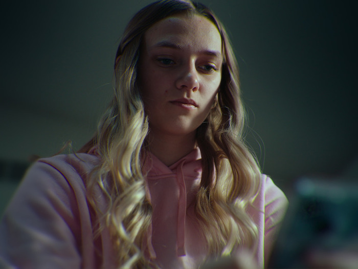 Teen girl with long blonde hair wearing pink hoodie, looking at phone, related to mom who catfished her own daughter case.