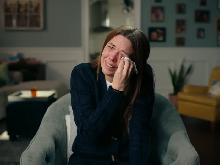 Woman wiping tears while sitting in armchair, related to expert explaining mom who catfished daughter using Lo nickname in threats
