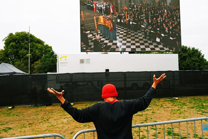 Person in a red hat raising arms watching a large screen showing a solemn event, capturing the atmosphere in London after Queen’s passing
