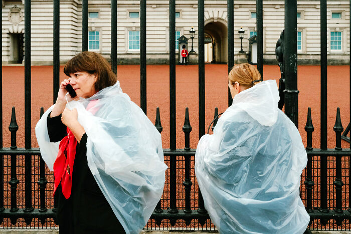 Two women in rain ponchos stand by iron gates in London, capturing the atmosphere after the Queen’s passing.
