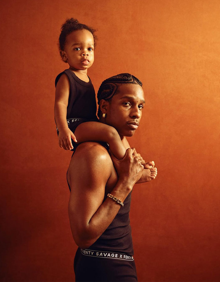 Man carrying toddler on shoulders wearing matching black Fenty Savage outfits in front of brown backdrop related to Rihanna fans urge cruel name.