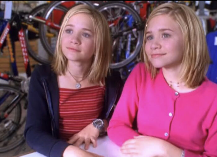 Young twin sisters Mary-Kate and Ashley Olsen sitting indoors with bicycles in the background, looking thoughtful. Young twin sisters Mary-Kate and Ashley Olsen sitting indoors with bicycles in the background, looking thoughtful.