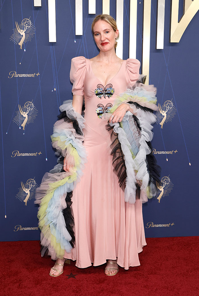 Woman in a pink dress with butterfly details and colorful tulle shawl posing on the red carpet at the 2025 Emmys fashion event.