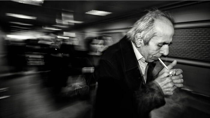 Black and white street photograph capturing an older man lighting a cigarette with blurred pedestrians in the background.