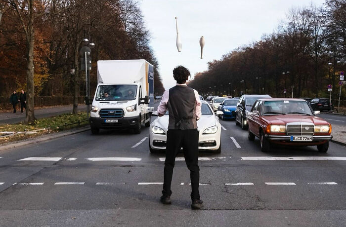 Street photograph of a juggler timing clubs perfectly in front of traffic, showcasing entertaining street photography moments.
