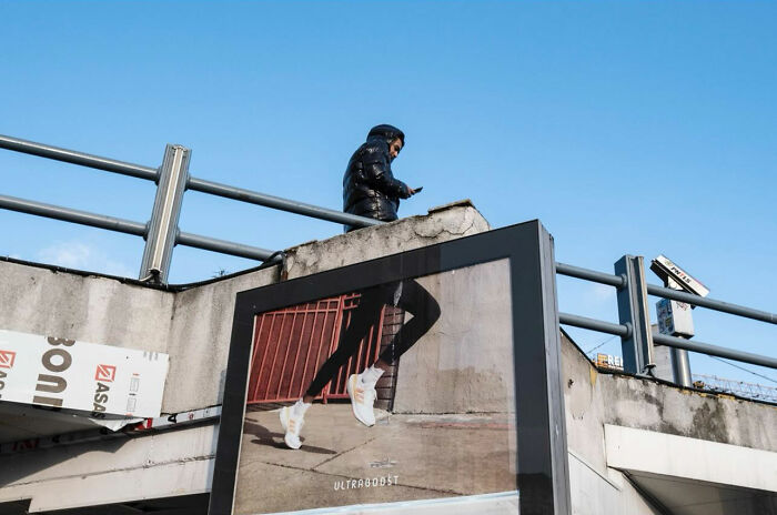 Man in black jacket walking above a street photograph billboard showing legs in running shoes, illustrating timing in street photography.